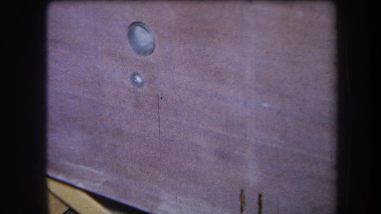 A 1964 Yucaipa home-movie frame shows two glossy circular drops on a pink-brown wooden surface with faded color and grain.