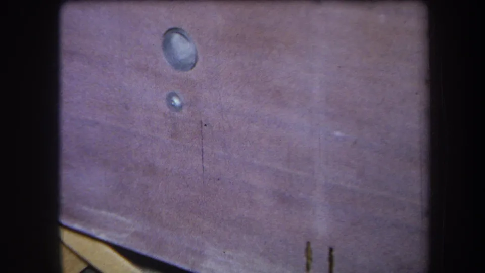 A 1964 Yucaipa home-movie frame shows two glossy circular drops on a pink-brown wooden surface with faded color and grain.