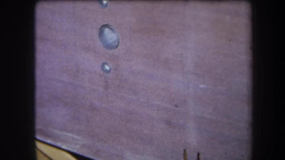 The 1964 8mm frame holds on two shiny drops on wood, with a pale edge entering at the lower left and visible film wear.
