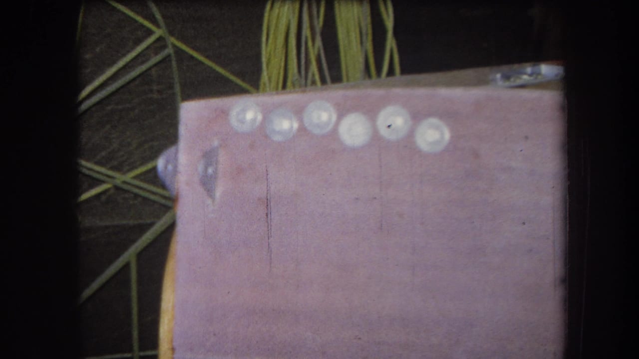 1964 Yucaipa-area 8mm frame showing a pink craft surface with pale round elements and green strands behind it.
