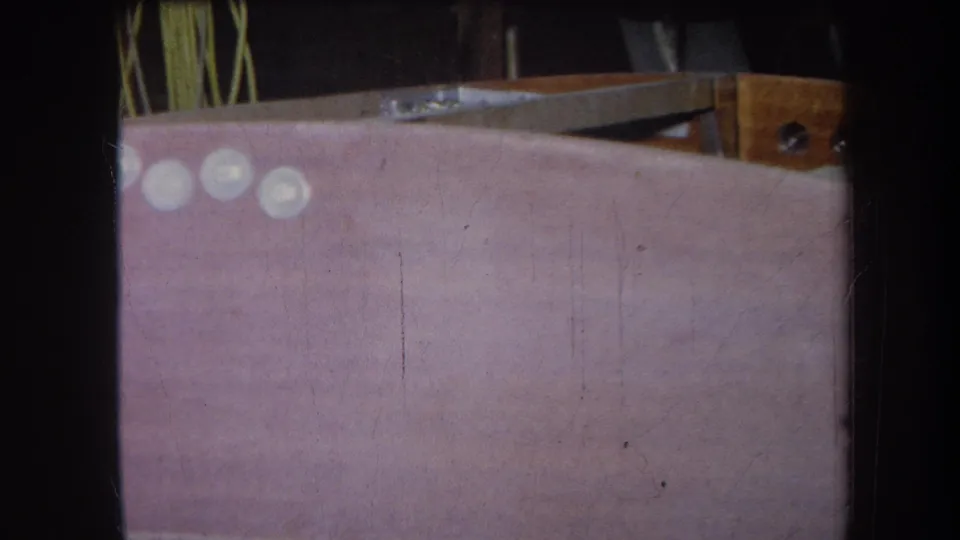 1964 Yucaipa-area 8mm frame of the pink craft surface with pale round pieces, green strands, and a wooden structure at right.