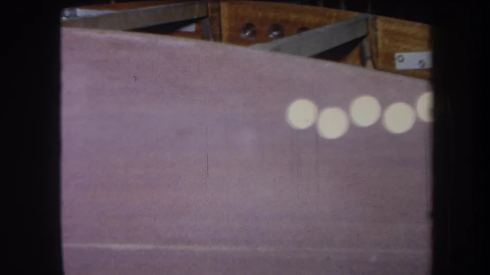 1964 8mm frame showing a purple-toned painted surface, wood hardware above it, and a row of white reflections.