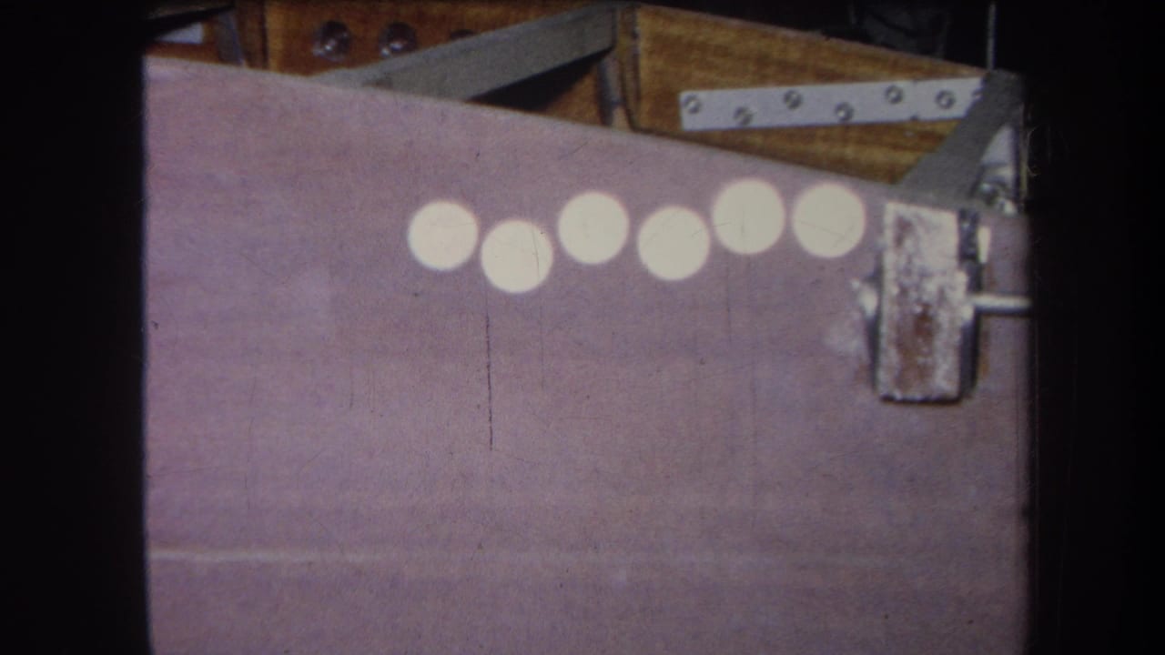 1964 8mm frame from Yucaipa showing six white circles across a purple-painted surface with rusty metal hardware and wood backing.