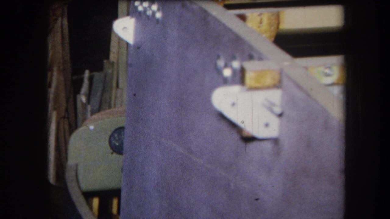 1964 8mm frame from Volmer Yucaipa showing a purple-painted wood structure with white brackets and stacked materials in back.