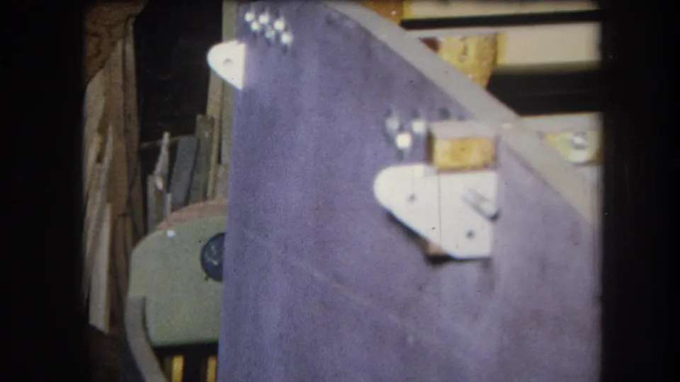 1964 8mm home-movie view of a purple-painted wooden structure and metal brackets in a cluttered California storage setting.