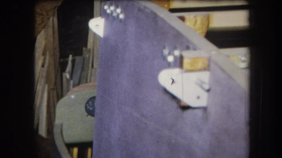 1964 8mm frame from Yucaipa showing a purple-painted wood panel with silver brackets, bolts, and surrounding workshop materials.
