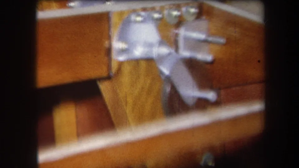 1964 8mm close-up showing hinge hardware, screws, and joined wooden boards, likely part of a furniture or workshop object.