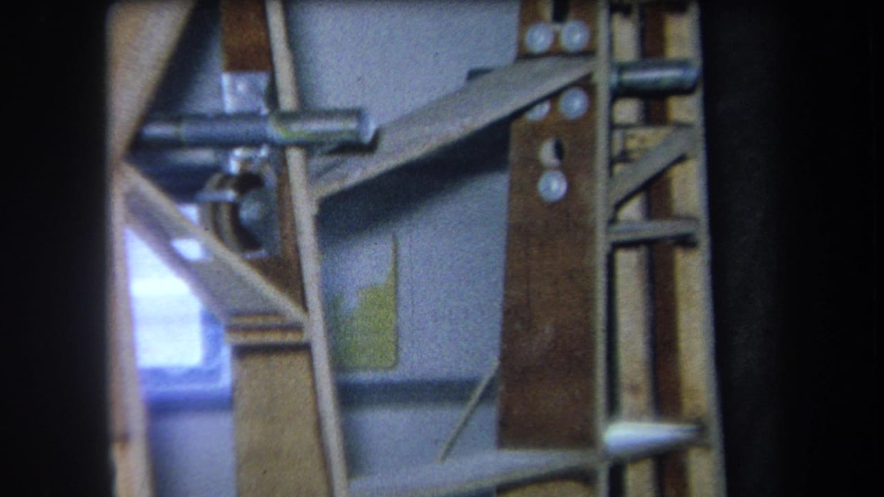 1964 Yucaipa home-movie frame showing wooden shelving, metal rods, angled braces, and a bright opening at left.