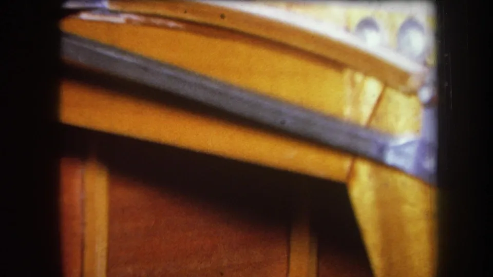 1964 8mm close-up in Volmer Yucaipa showing varnished wooden furniture and a dark metal strip in dim storage light.