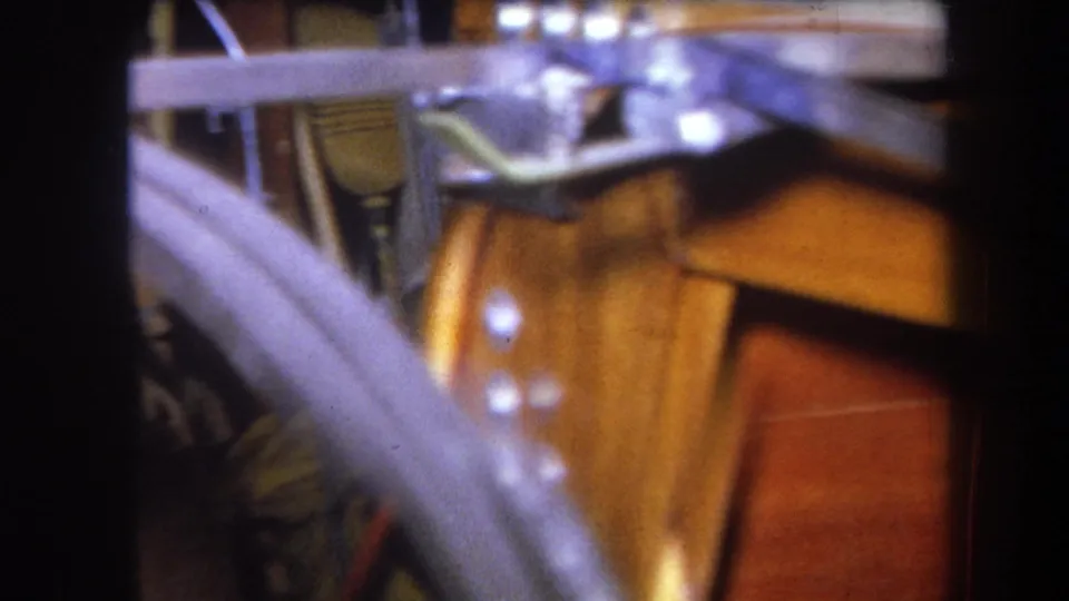 1964 Volmer Yucaipa storage close-up with pale metal tubing crossing wood, brass-toned hardware, and small bright fasteners.