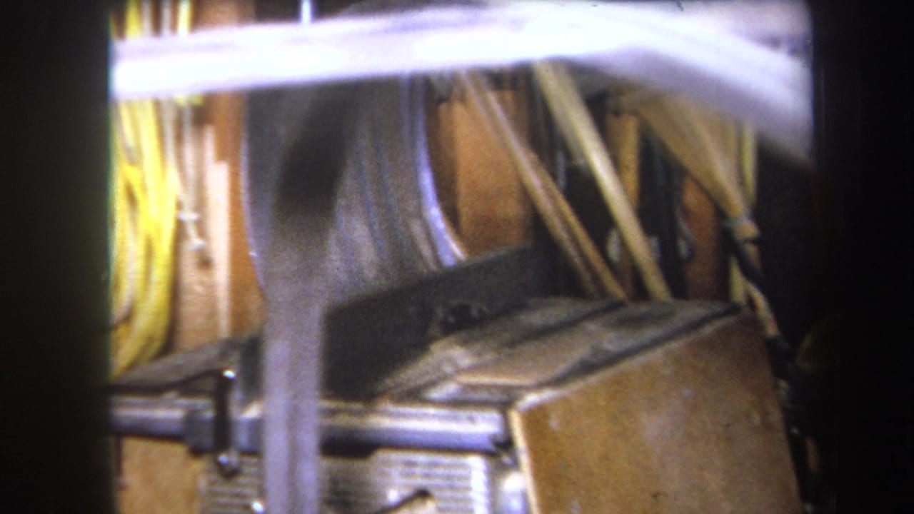 1964 8mm frame from Volmer Yucaipa showing wooden boards, a dark case or tool, and metal hardware in a dim workshop interior.