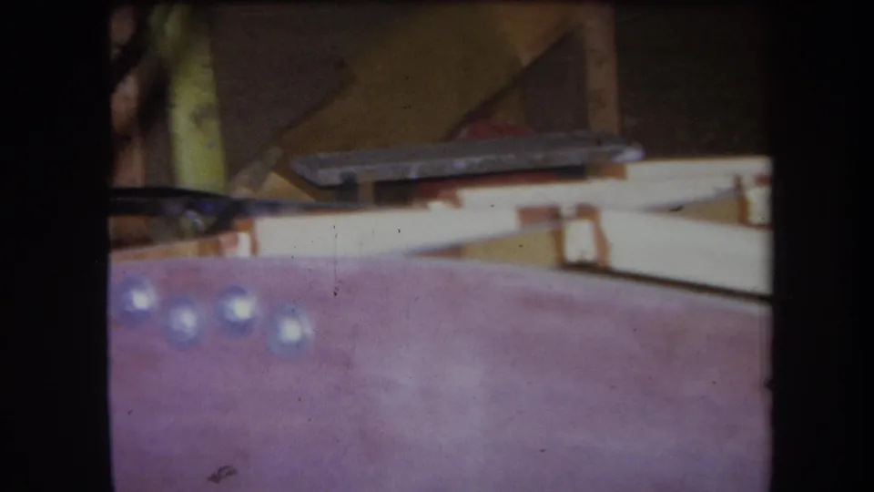 Soft 1964 8mm workshop frame showing a purple-pink foreground surface, pale dots, wood pieces, and a small metal fitting.