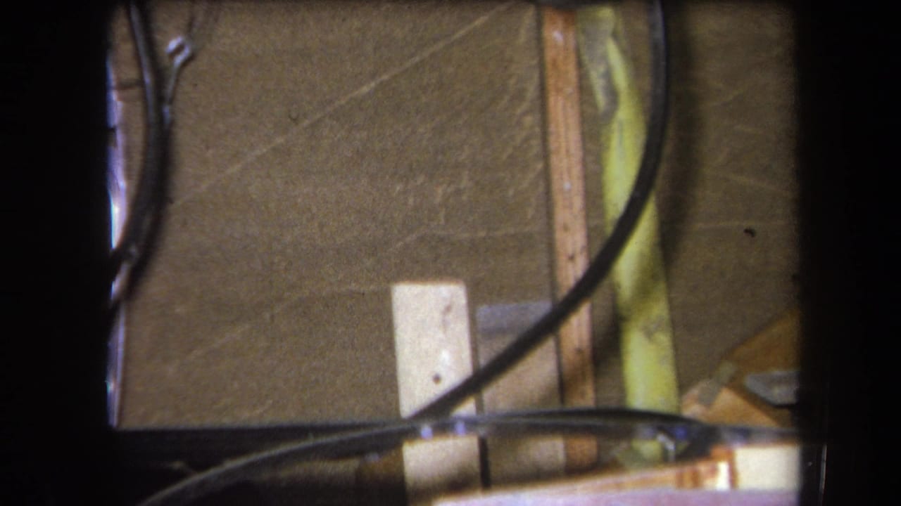 1964 Yucaipa 8mm frame showing black tubing, thin wires, wood strips, and a pale rectangular block against a brown textured wall.