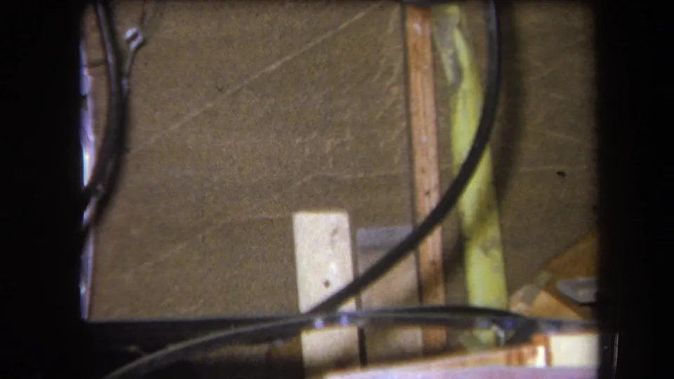 1964 Yucaipa 8mm frame showing black tubing, thin wires, wood strips, and a pale rectangular block against a brown textured wall.