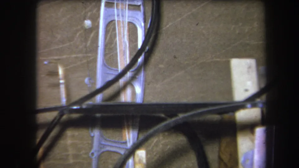 1964 Yucaipa 8mm frame showing crossed black tubing, thin wires, a translucent plastic form, and a pale block on a brown wall.