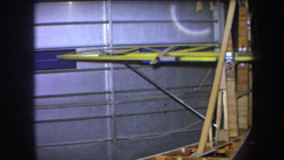A later frame in the 1964 home movie shows the yellow aircraft wing section and wooden tail framing from a close indoor angle.