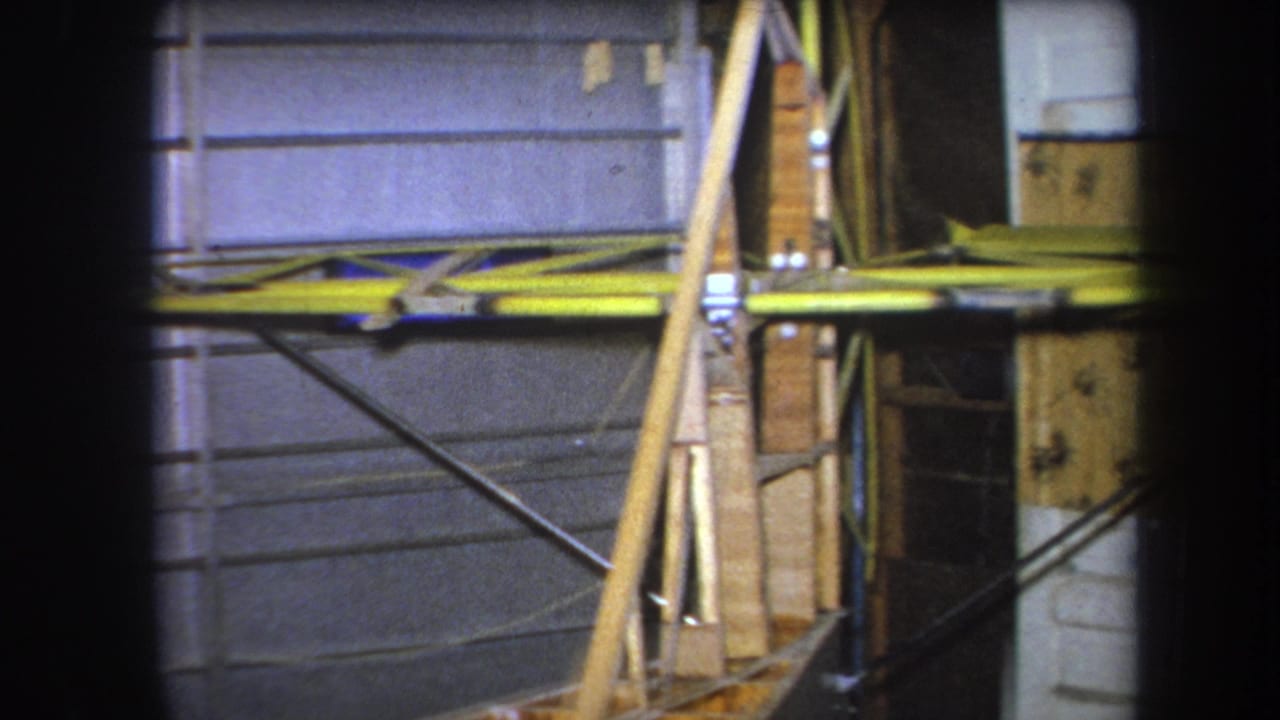 Close home-movie view of wood and yellow metal bracing set against gray wall panels in the 1964 California construction footage.