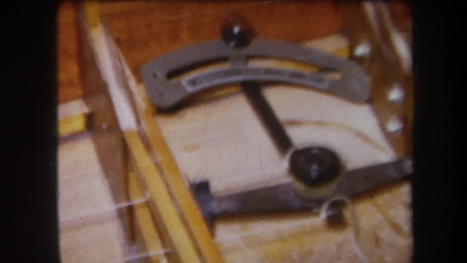 1964 8mm close-up from Yucaipa showing a wooden stringed instrument frame with a curved metal lever, tuning hardware, and dark adjustment knobs.