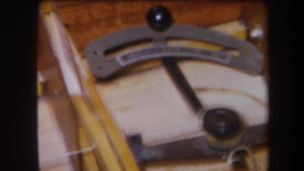 1964 8mm frame showing the stringed instrument mechanism from a slightly shifted angle, with the lever, knob, and wood grain in soft focus.