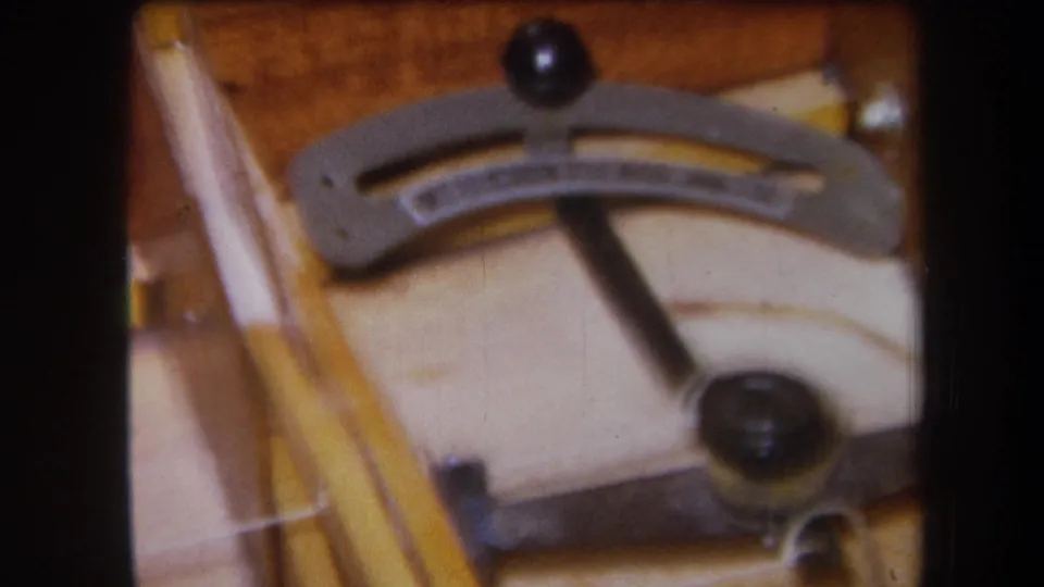 1964 8mm frame showing the stringed instrument mechanism from a slightly shifted angle, with the lever, knob, and wood grain in soft focus.