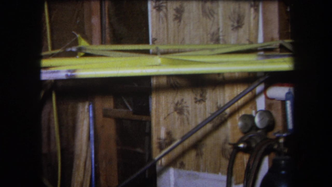 1964 Yucaipa home-movie frame showing a workshop interior with exposed wood, patterned wall covering, yellow beam, and gas cylinder gauges.