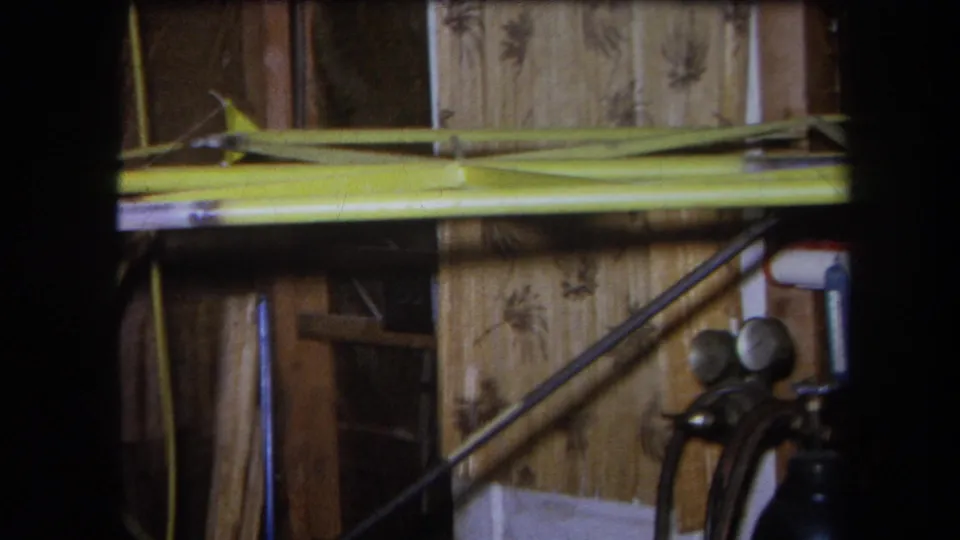 1964 Yucaipa home-movie frame showing a workshop interior with exposed wood, patterned wall covering, yellow beam, and gas cylinder gauges.