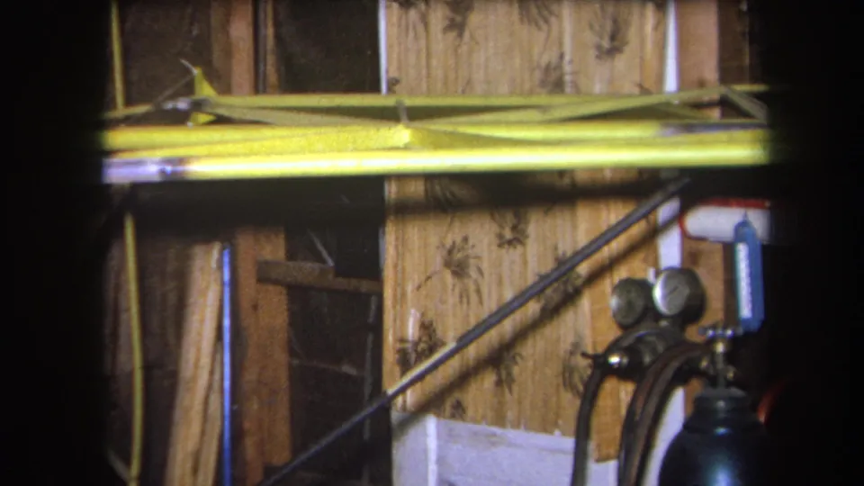 8mm frame of a private workshop interior in Yucaipa, showing exposed wood, a yellow beam, diagonal supports, and a green cylinder with gauges.