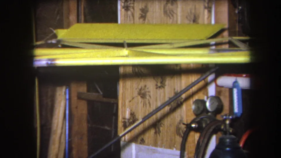Final 1964 home-movie frame of a Yucaipa workshop, with a yellow frame or beam, wall panels, diagonal rods, and gas cylinder gauges.