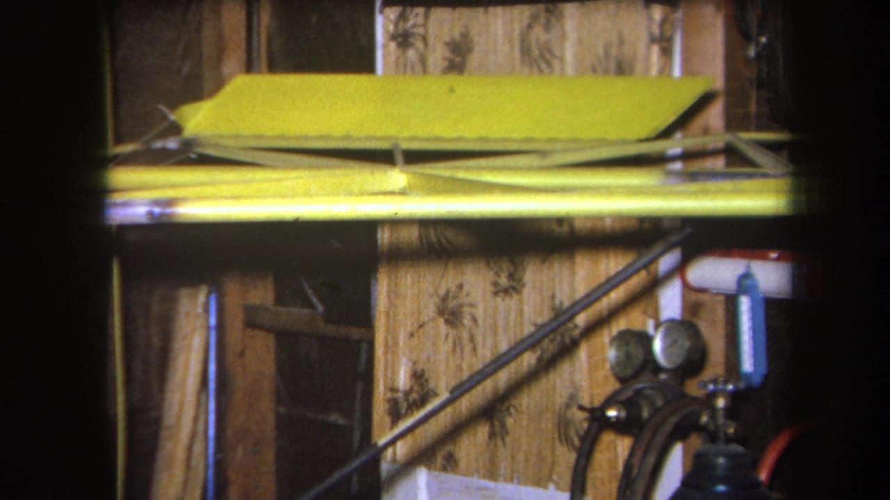 1964 Yucaipa home-movie frame showing a yellow model aircraft part in a dim garage or workshop, with wood paneling and gauges in the background.