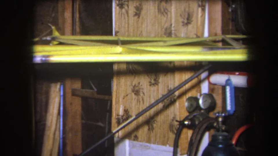 The 8mm frame shows the yellow model aircraft component in a private Yucaipa workshop, with floral wood paneling and partial equipment visible.