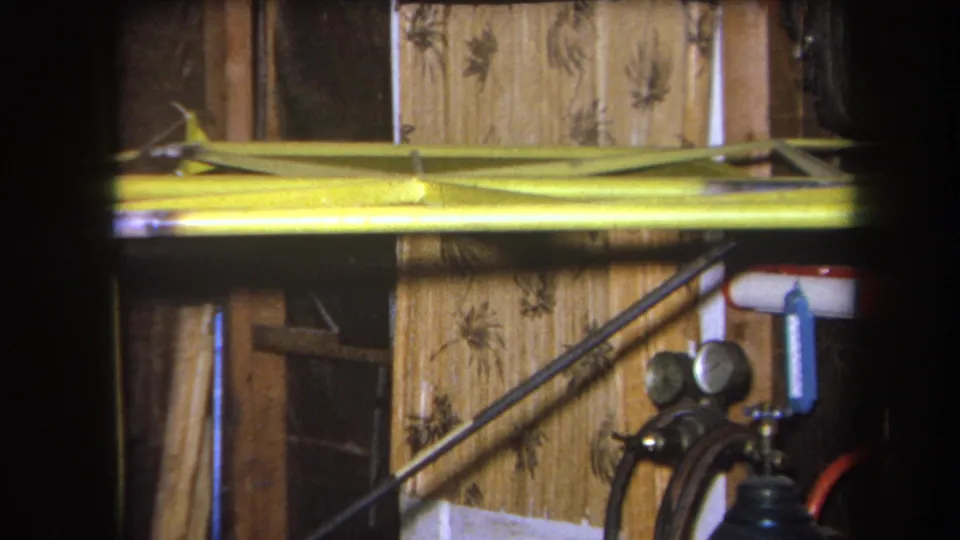 Final 1964 Yucaipa frame of the yellow model airplane part in a dim garage or workshop, with paneling, rod, and gauge equipment visible.