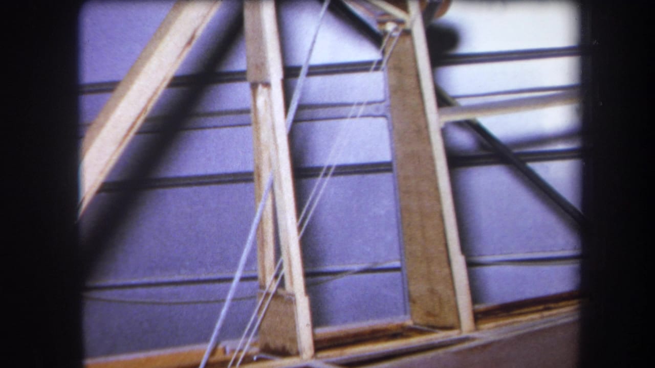 1964 Yucaipa home-movie frame showing wooden bracing, white cables, and metal rods in a warehouse-like interior.