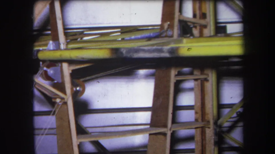 1964 8mm construction-site detail with timber uprights, a yellow horizontal beam, and dark rods against pale panels.
