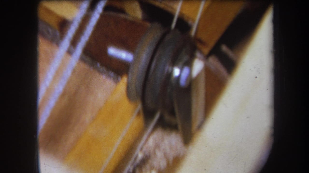 1964 Yucaipa-area 8mm close-up of a dark pulley and pale thread running across worn sewing-machine components.