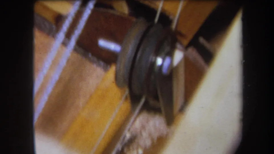 1964 8mm frame showing a sewing-machine pulley and thread path against warm wood-toned and metal surfaces.