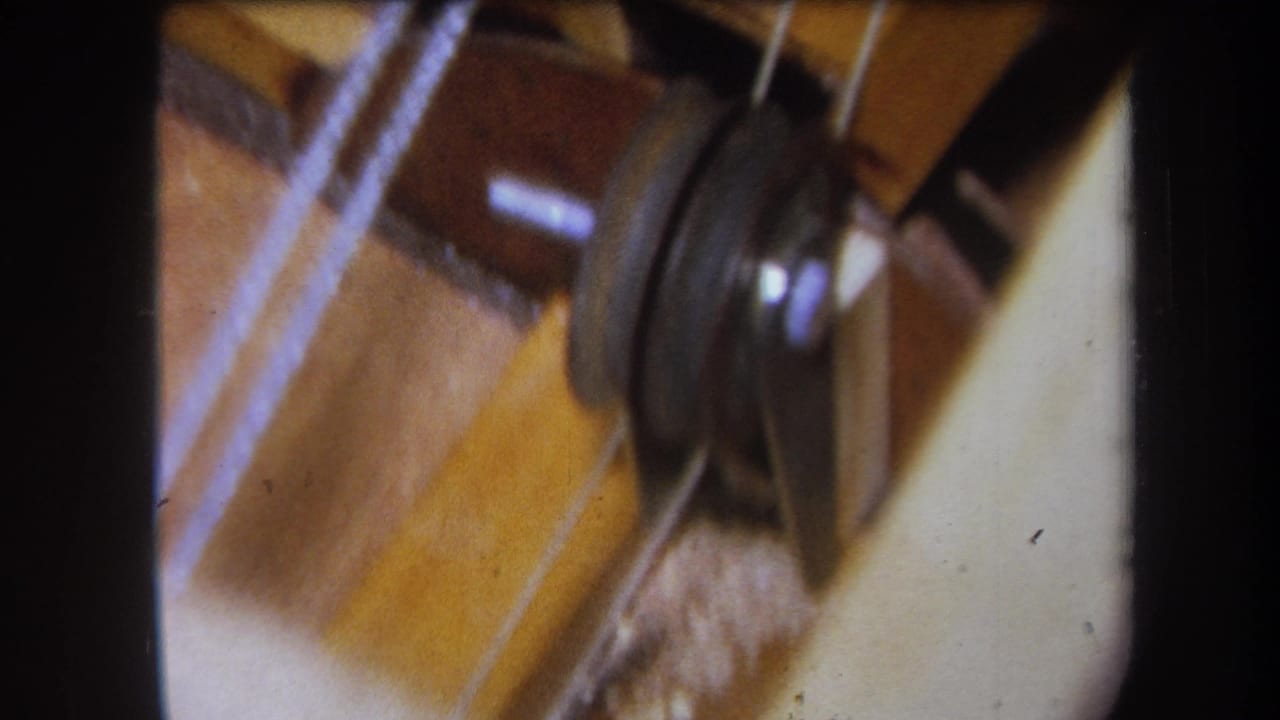 1964 Yucaipa home-movie frame showing a close view of a wheel, cords, and wooden parts on a homemade workshop mechanism.