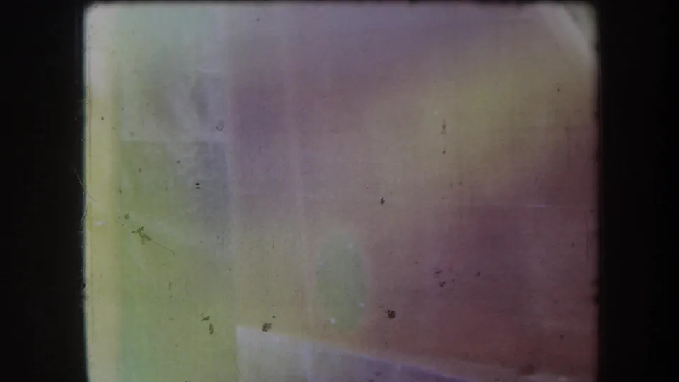 Faded 8mm frame with pale green and pink color wash, dust specks, and dark film edges near the end of the shot.