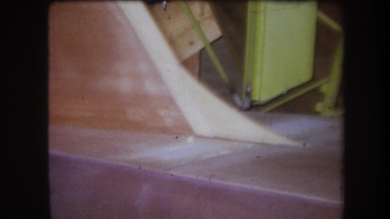 1964 Yucaipa 8mm close-up of an old agricultural machine section with a pale angled part and yellow-green frame above a reddish surface.