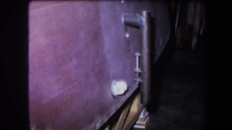 1964 Yucaipa 8mm close-up of old agricultural equipment, showing a worn side panel, vertical metal fitting, pale knob, and shadowed interior edge.