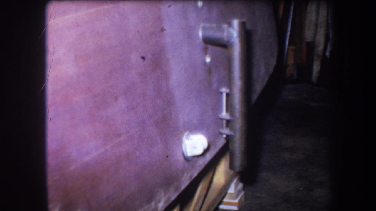 1964 8mm frame from Yucaipa showing a close-up of an old boiler side, vertical pipe, handle and white circular fitting in a dim utility space.