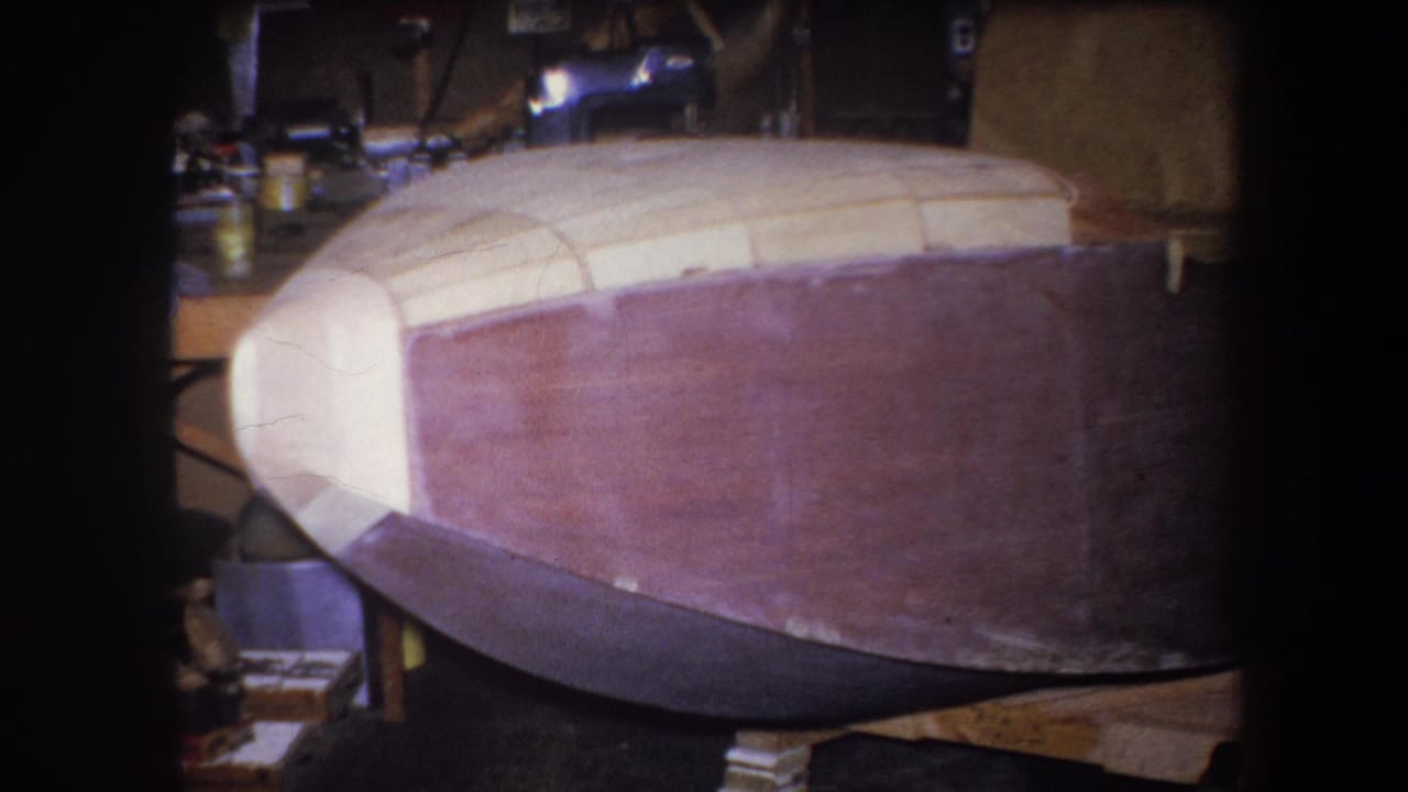 1964 Volmer Yucaipa 8mm frame showing an unfinished wooden boat hull with pale bow and reddish side panels in a private workshop.