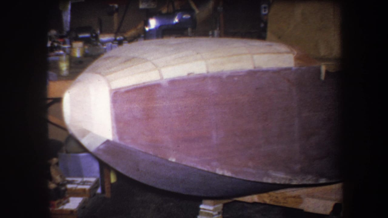 1964 Volmer Yucaipa frame of the same unfinished hull, showing the pale upper structure, reddish side, and dim garage background.
