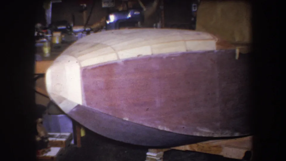 Side view of an unfinished wooden boat hull in the 1964 Volmer Yucaipa home-movie footage, with supports and shop clutter around it.