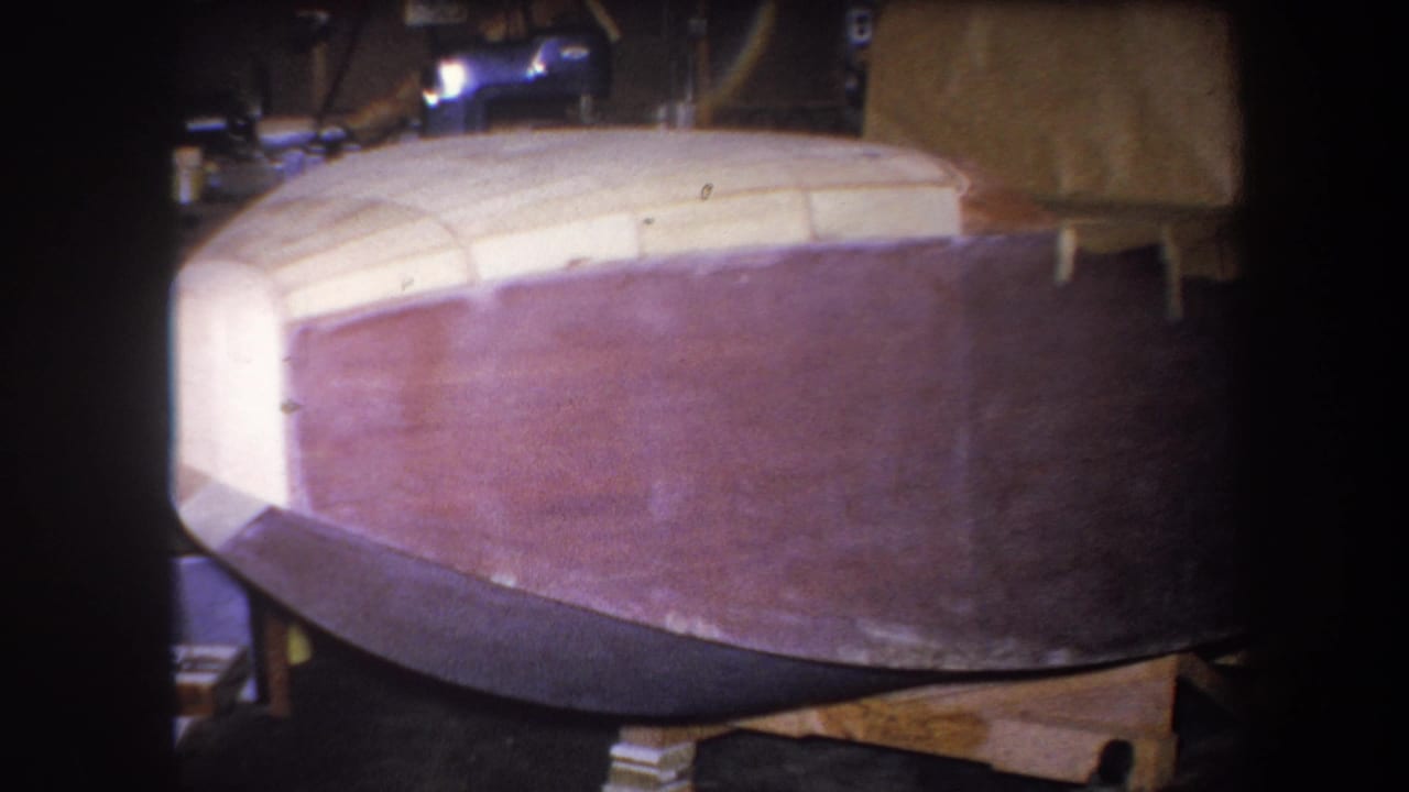 Brown wooden boat hull on a workshop support frame.