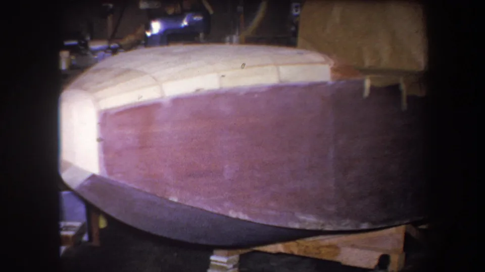 1964 Yucaipa home-movie frame showing a brown wooden boat hull with pale framing in a dim workshop.