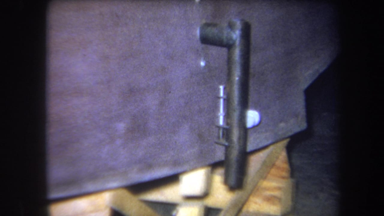 Close 1964 home-movie view of a painted wooden piece held above pale blocks with black metal hardware attached.
