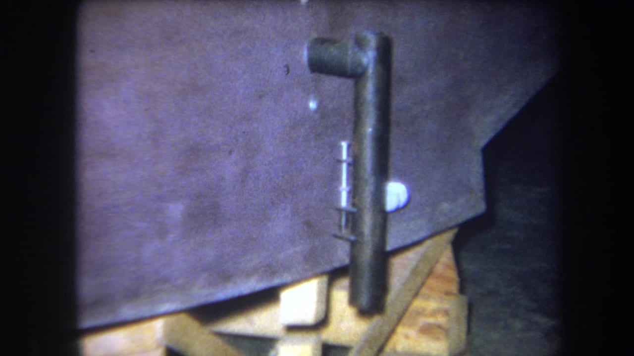 1964 8mm frame from Yucaipa showing a close workshop view of a metal pipe, wooden blocking, and part of a boat construction setup.