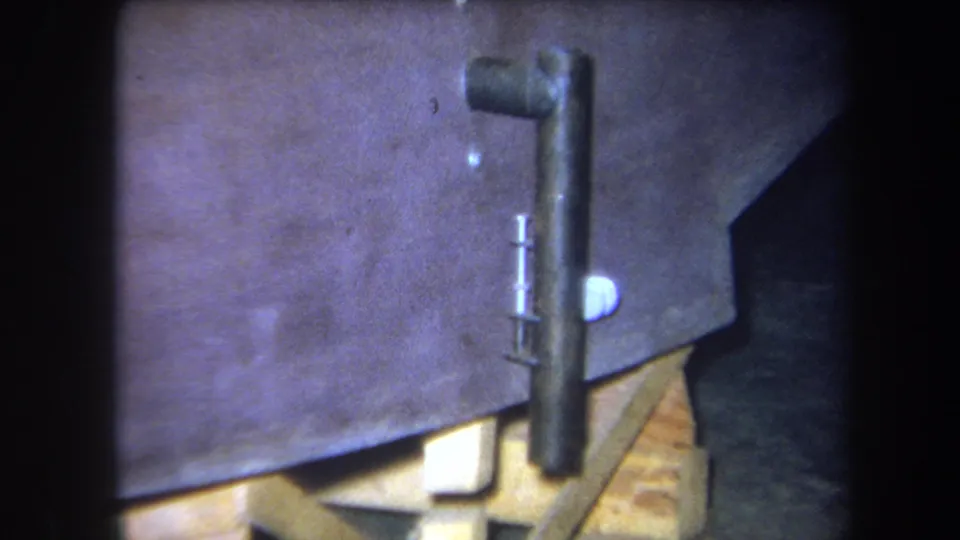 1964 8mm frame from Yucaipa showing a close workshop view of a metal pipe, wooden blocking, and part of a boat construction setup.