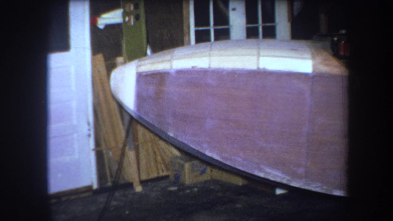 1964 8mm frame from Yucaipa showing the front section of a curved boat hull under construction in an indoor workshop.
