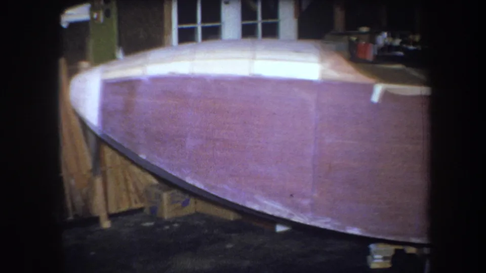 1964 8mm frame from Yucaipa showing a partially finished boat hull, stacked boards, and workshop windows in the background.